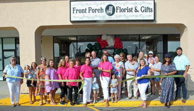 130807082_web1_Ribbon-cutting
Emma Smith uses a large pair of scissors to cut a ceremonial ribbon Friday morning, Aug. 3, to celebrate the grand re-opening of Front Porch Florist & Gifts. She and her parents, Jennifer Barwick, standing next to her in the pink shirt holding the ribbon, and Ronald Barwick, standing behind and between the two, are the new owners of the business. Smith is the granddaughter of Betty Sue Rose, in the colorful floral design shirt standing next to her, and the Rev. Paul Rose, standing next to his wife and wearing a blue shirt and holding a child. At left is Julie Beck, Mount Olive Area Chamber of Commerce president. (Steve Herring|mountolivetribune.com)