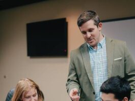 
			
				                                Assistant Dean of the Tillman School of Business Waylon Woodall (standing) supervises students with the VITA tax preparation process. (UMO photo)
 
			
		